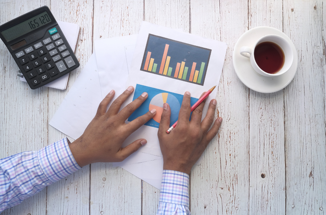 Cenital plane of human hands touching a paper with statistics content, with a cup of coffee on the right side and a calculator on the left side.