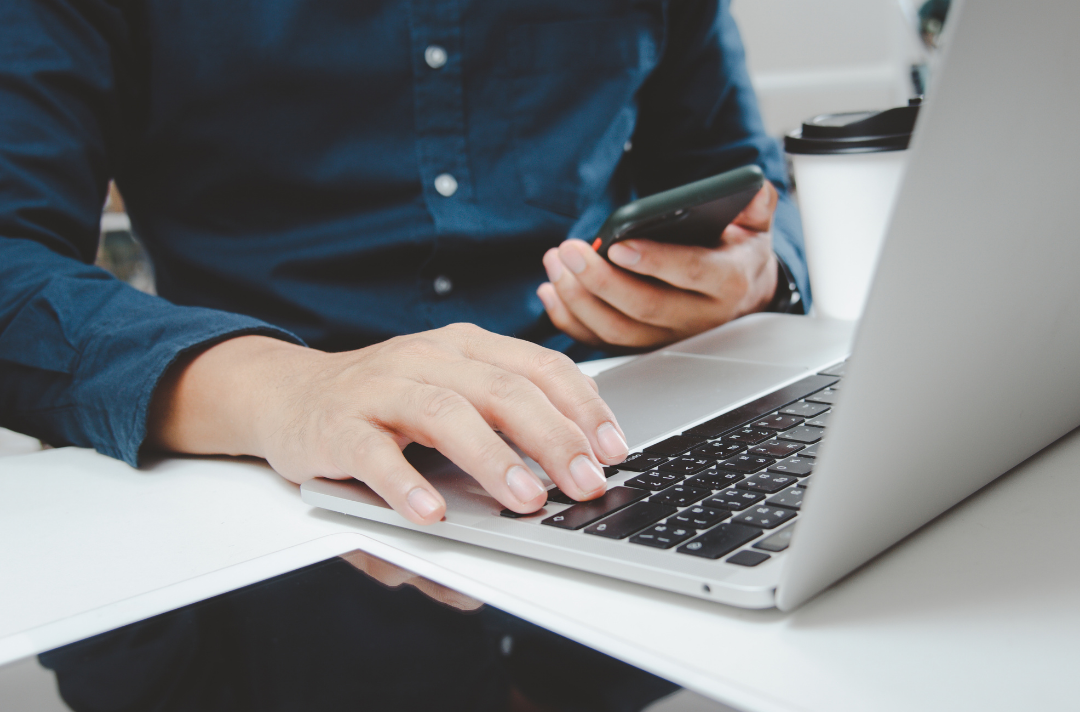 An image of the foreground of a man using a computer with his right hand holding a smartphone with his left one.